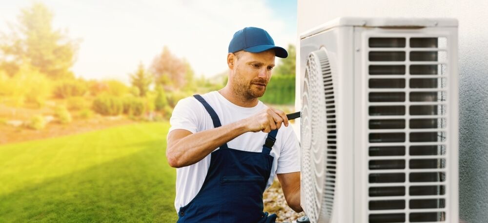 A technician in blue overalls and a cap uses a screwdriver to repair or install an outdoor air conditioning unit next to a grassy yard.