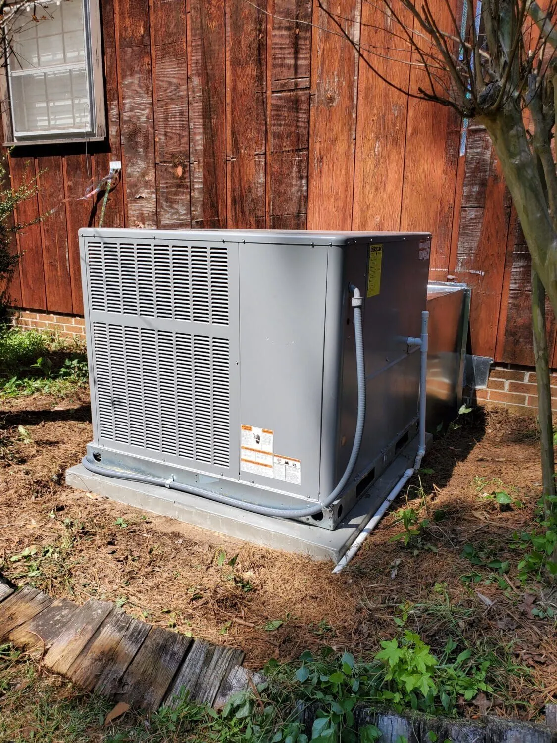 An outdoor HVAC unit installed next to a wooden building, with a dirt and grassy area surrounding it.