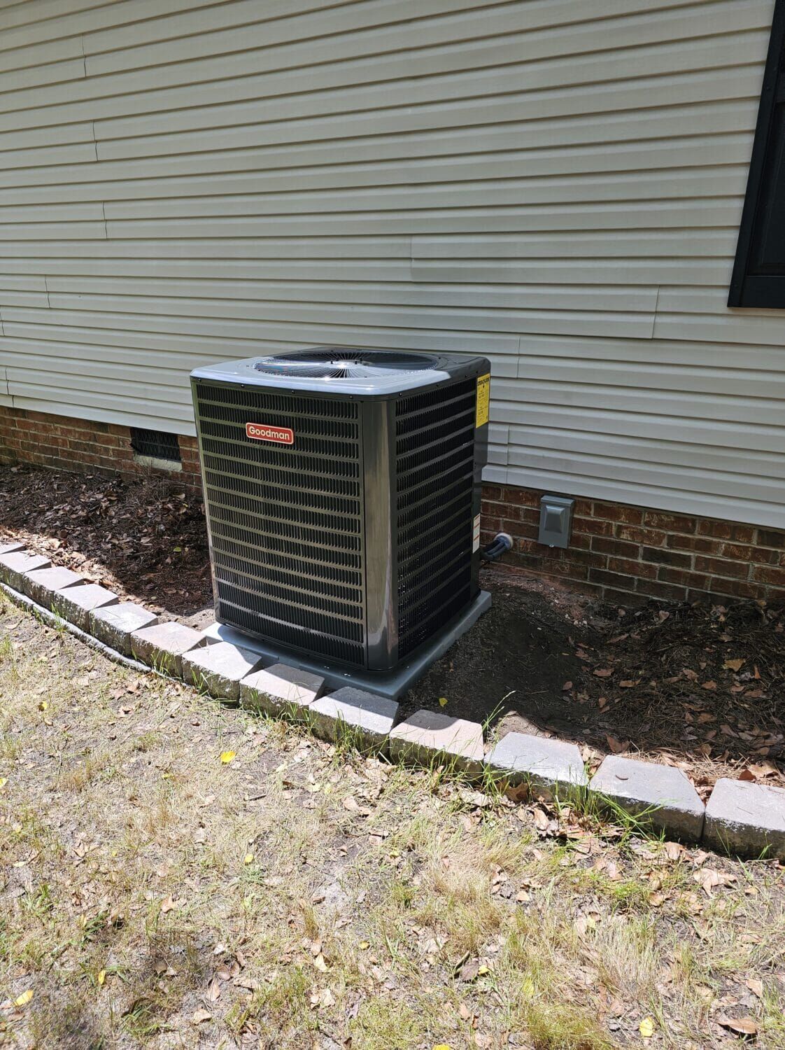 An outdoor air conditioning unit placed on a concrete pad beside a house with tan siding and a surrounding mulch bed.