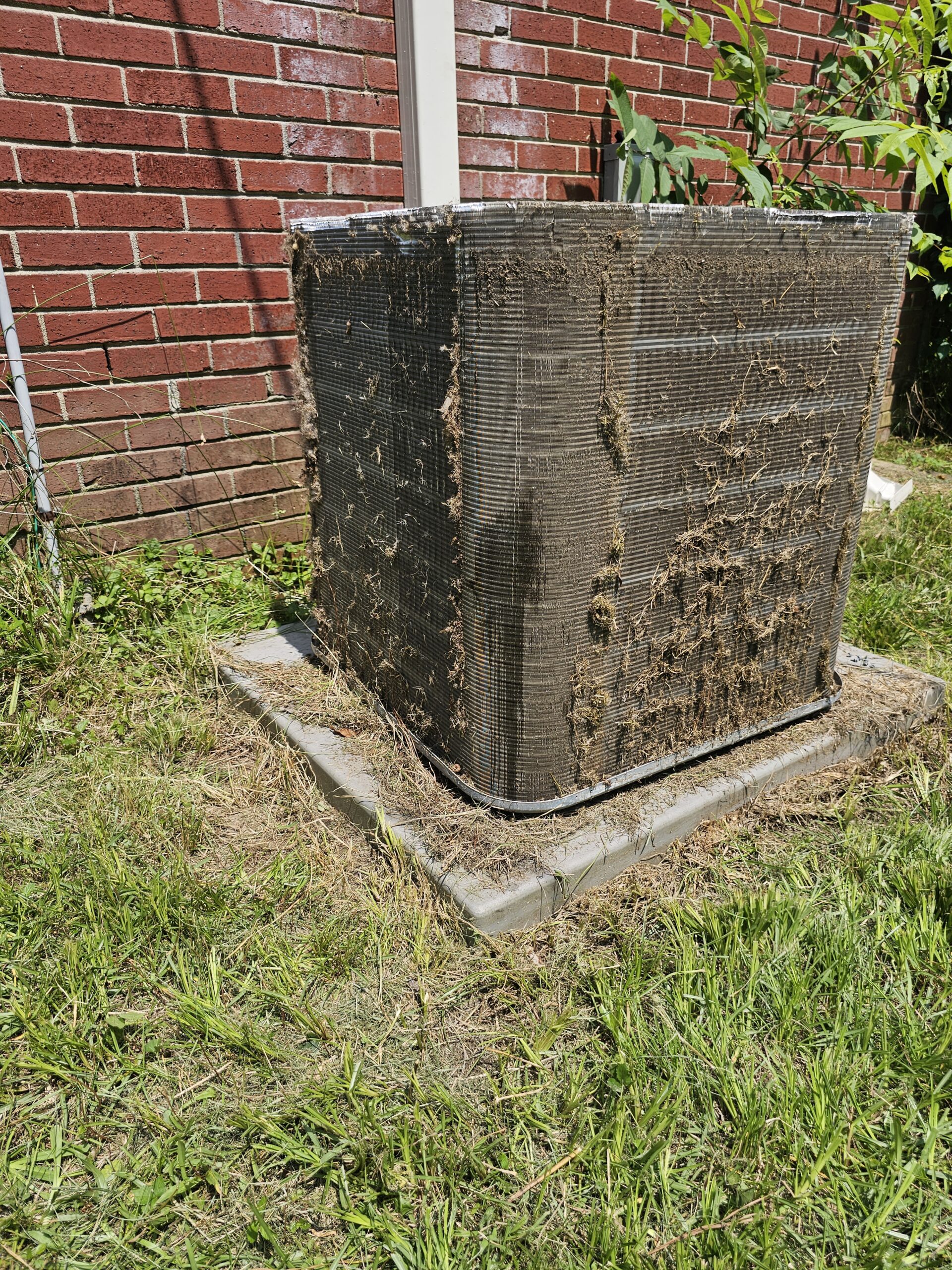 An outdoor air conditioning unit with dirty and debris-covered coils sits on a concrete pad next to a brick building in a grassy yard.