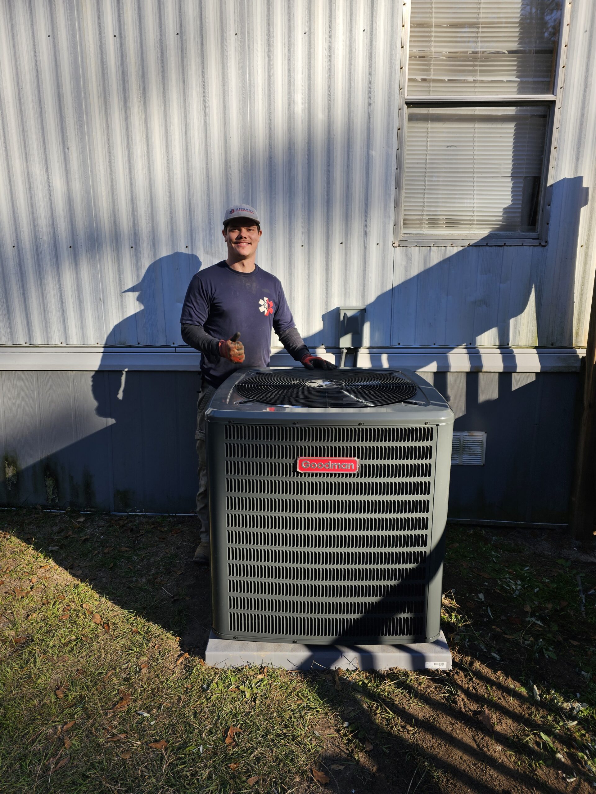 A person stands outdoors next to a Goodman air conditioning unit installed beside a building, holding a tool and smiling at the camera.
