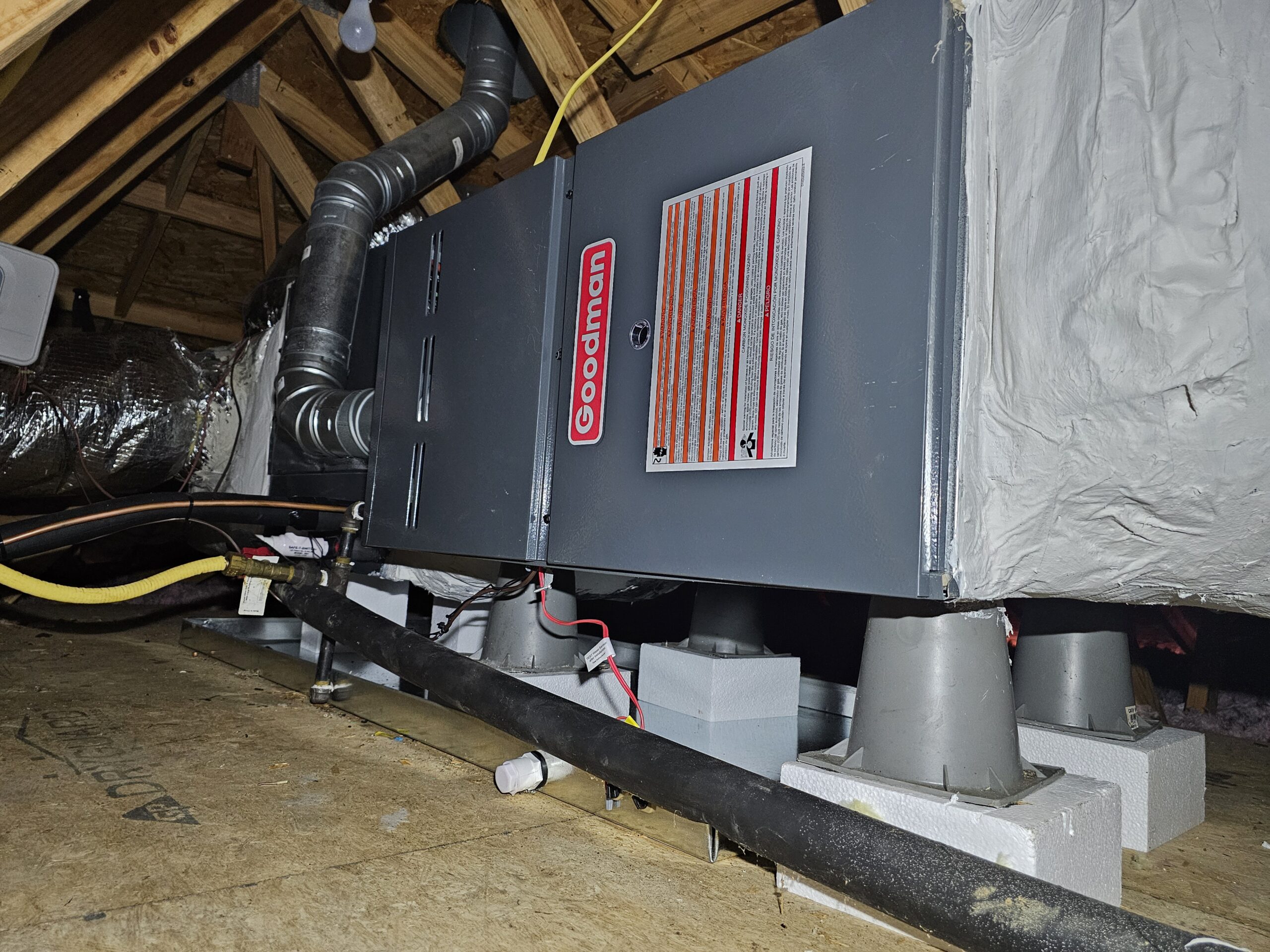 A Goodman HVAC unit is installed in an attic, supported by concrete blocks, with ducts and wiring visible.