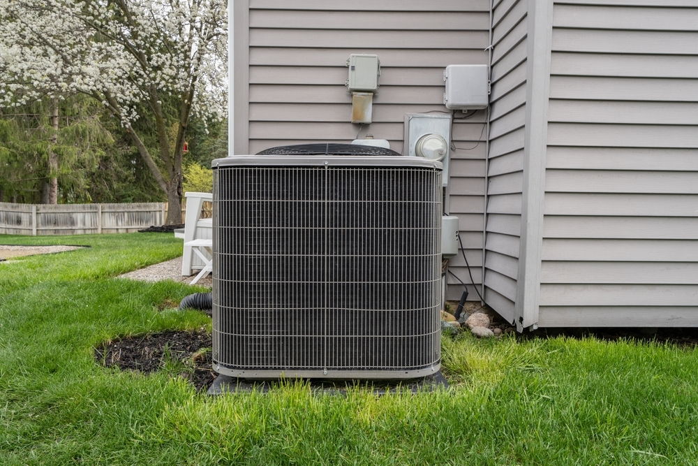 Outdoor air conditioning unit beside a gray house, surrounded by green grass and a small garden.