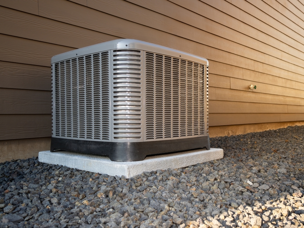 An outdoor air conditioner unit sits on a concrete pad, surrounded by gravel, against the wall of a building with horizontal siding.