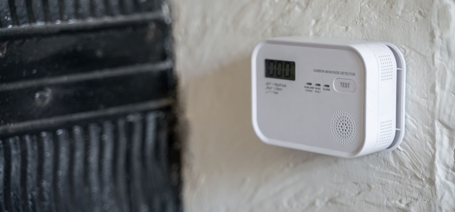 A white carbon monoxide detector with a digital display and a test button is mounted on a textured white wall next to a black door frame.