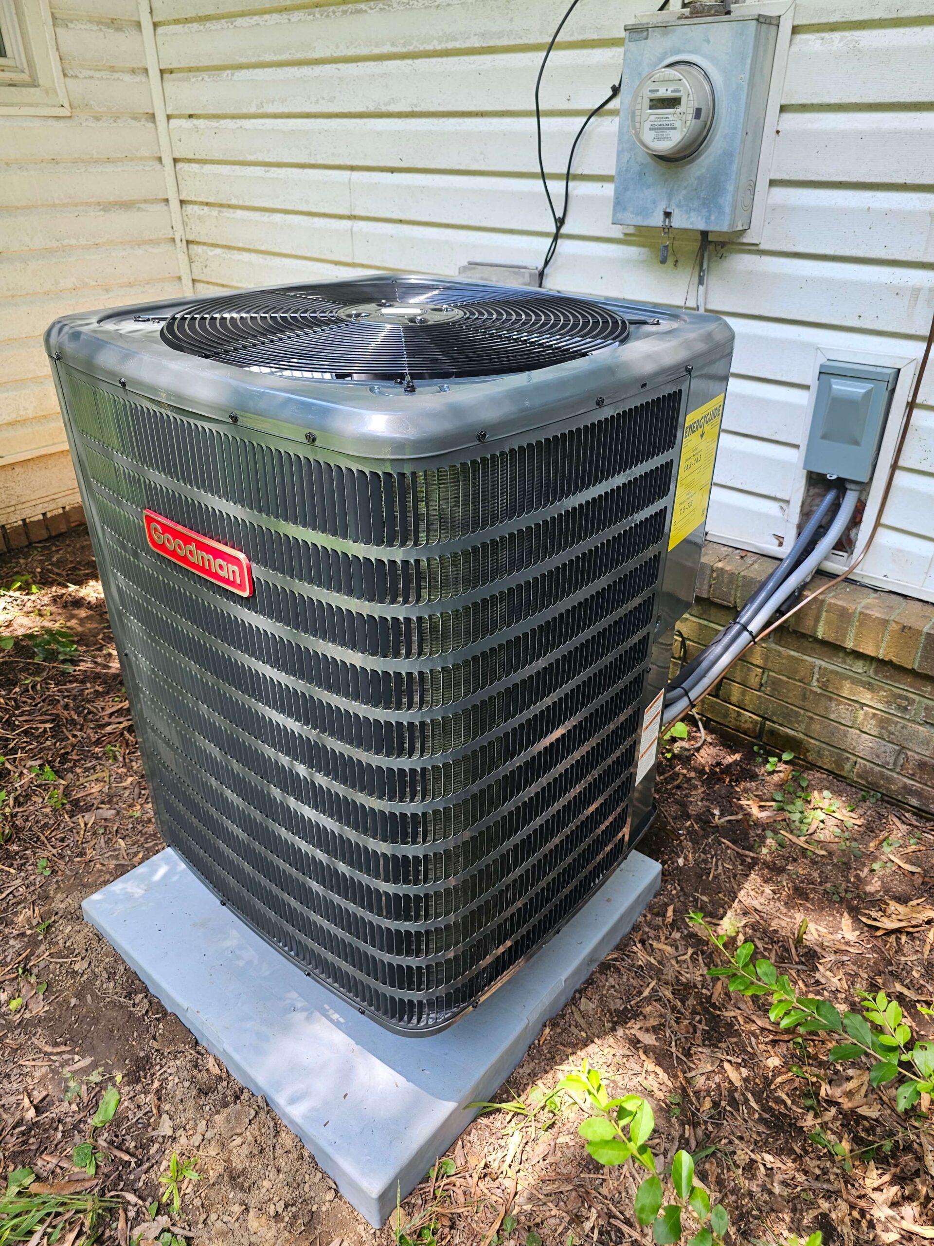 A Goodman outdoor air conditioning unit sits on a concrete pad next to a house, with electrical wiring connected to a nearby meter and junction box.