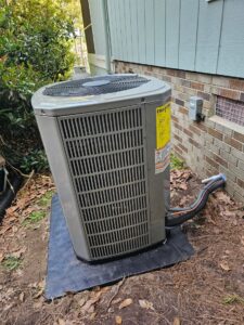 Outdoor air conditioning unit situated on a black pad next to a brick and wood-sided house, with pipes connected and some fallen leaves on the ground.