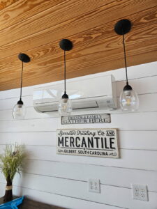 Three pendant lights hang from a wooden ceiling. Below them is a wall-mounted air conditioner and two decorative signs, with a vase of flowers on a ledge.