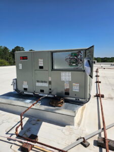 A commercial HVAC unit with an open panel is mounted on a flat rooftop. Tools and piping are visible nearby under a clear blue sky.