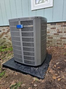 An outdoor air conditioning unit sits on a concrete pad next to a house with blue siding and brickwork near the foundation.
