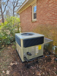 A rectangular air conditioning unit is installed outside a brick house, surrounded by grass, leaves, and shrubs.