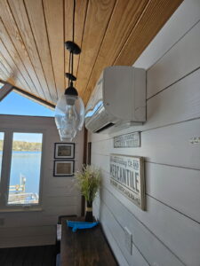 Interior of a room with wooden ceiling, pendant lights, an air conditioner, framed pictures, and decorative signs on white paneled walls. A window offers a view of a lake or river.
