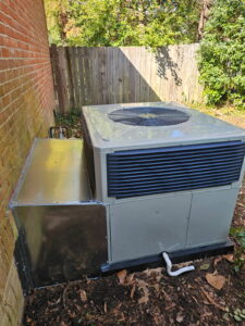 Air conditioning unit installed outdoors next to a brick wall and wooden fence, with surrounding greenery.
