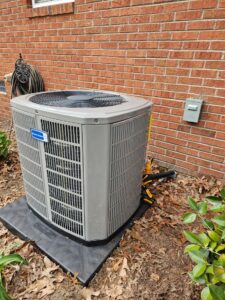 Outdoor air conditioning unit installed next to a red brick wall, surrounded by fallen leaves and foliage, with a hose coiled nearby.