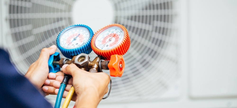 Person using a manifold gauge set to check pressures on HVAC equipment, with a blurred fan unit in the background.