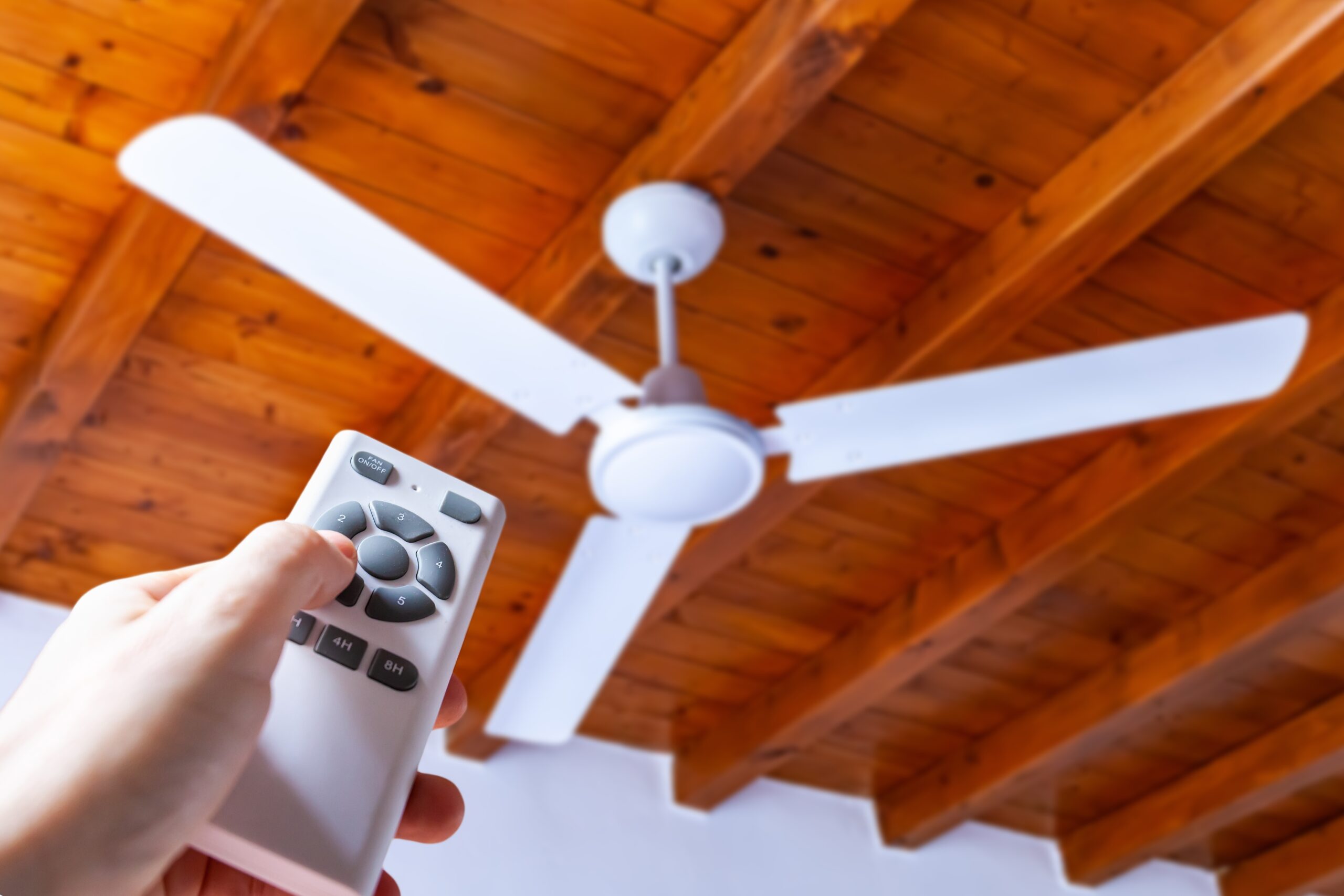 A hand holds a remote control pointed at a white ceiling fan mounted on a wooden ceiling.