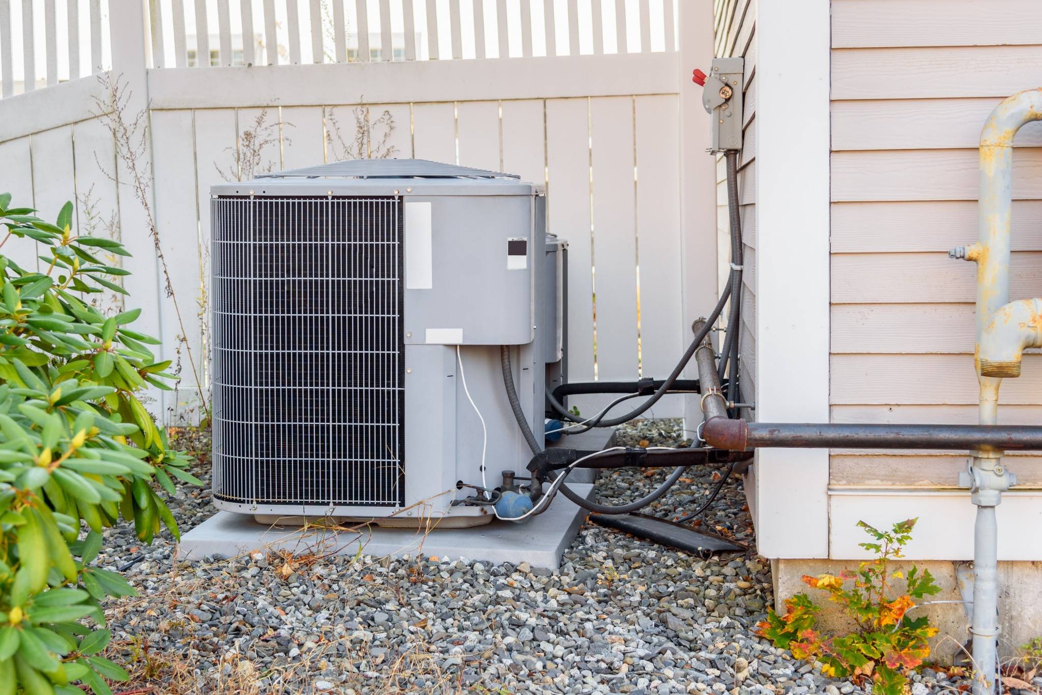 Outdoor air conditioning unit next to a house, connected with cables and pipes, surrounded by gravel and some plants near a white fence.